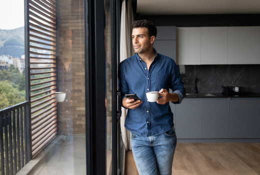 Eine Person, steht in einer modernen Küche und hält eine Tasse Kaffee in der einen Hand und ein Smartphone in der anderen. Im Hintergrund sind große Fenster mit Blick auf eine Landschaft mit Bäumen und Bergen zu sehen.